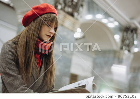 Beautiful Caucasian woman wearing coat, beret, scarfsitting alone on Saint Petersburg metro station reading a book. Image with selective focus and noise effect 90752102