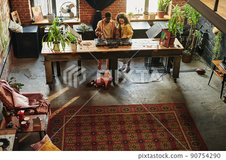 Young interracial couple in casualwear standing by long wooden table with dj set Young interracial couple in casualwear standing by long wooden table with dj set 90754290