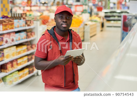 African American guy in red uniform and cap using digital tablet in supermarket 90754543
