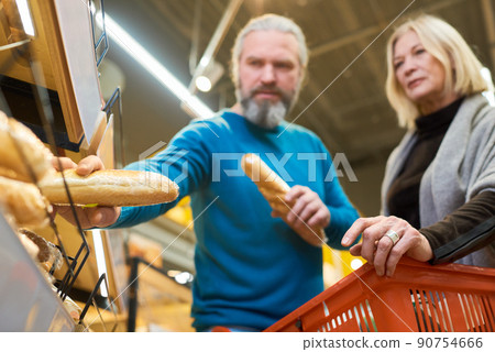 Mature male customer taking two fresh wheat baguettes from shelf in supermarket Mature male customer taking two fresh wheat baguettes from shelf in supermarket 90754666