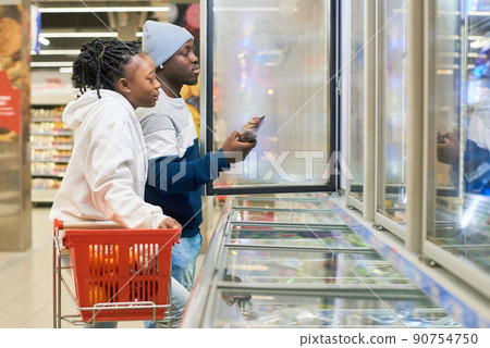 Young couple with shopping cart choosing frozen food products in supermarket 90754750