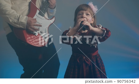 Little girl on stage in vintage dress, she sings into microphone and dances, her father plays an electric guitar. Color music is shining and smoke is billowing. Family time, creativity and hobbies 90756342