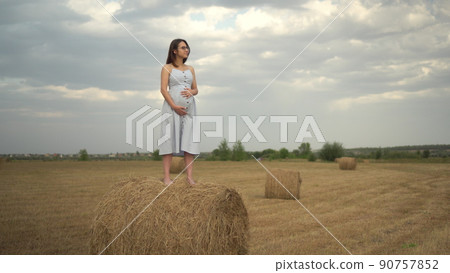 A young pregnant woman stands on a haystack in a field and holds her belly with her hands. Girl with glasses and a blue dress. A young pregnant woman stands on a haystack in a field and holds her belly with her hands. Girl with glasses and a blue dress. 90757852