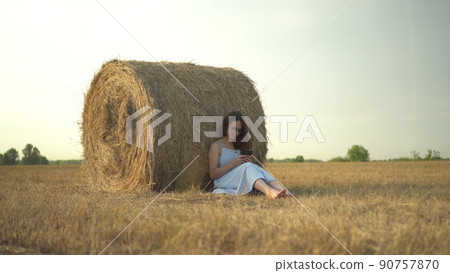 A young woman sits near a haystack in a field with a phone in her hands. A girl with glasses and a blue dress is texting on the phone. A young woman sits near a haystack in a field with a phone in her hands. A girl with glasses and a blue dress is texting on the phone. 90757870
