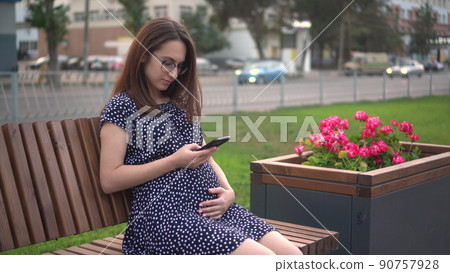 Young pregnant girl with a phone in her hands. A girl with glasses and a dress is texting on the phone. 90757928
