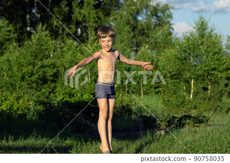 Happy smiling boy in wet shorts plays with water sprinkler in garden. 90758035