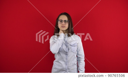 A young woman with glasses has a pain in her neck, she holds her neck with her hand. Shooting on a red background. A young woman with glasses has a pain in her neck, she holds her neck with her hand. Shooting on a red background. 90765506
