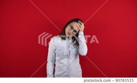 A young woman with glasses has a headache and holds it in her hands. Shooting on a red background. 90765515