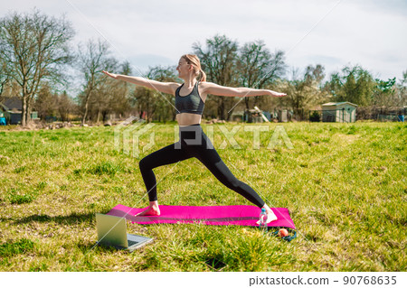 Smiling relaxed young fitness girl using laptop with online trainer, doing yoga at the park. Spring season sunny lawn mowing in the garden. 90768635