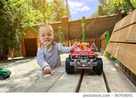 A little cute boy of one and a half years plays with toy car at the playground. Adorable toddler playing with cars and toys outdoor. Family, holiday, kids lifestyle concept. Selective focus A little cute boy of one and a half years plays with toy car at the playground. Adorable toddler playing with cars and toys outdoor. Family, holiday, kids lifestyle concept. Selective focus 90769091