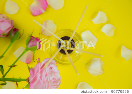A glass jar with scented oil and wooden incense sticks in the room, next to roses and rose petals on a bright yellow background. High quality photo A glass jar with scented oil and wooden incense sticks in the room, next to roses and rose petals on a bright yellow background. High quality photo 90770936