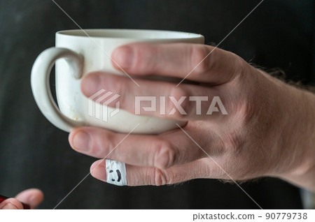 A man is holding a ceramic cup in his hand. The first phalanx of his little finger is wrapped in white ribbon. A smiling face is painted on the bandage. Positive emotion concept. Selective Focus. A man is holding a ceramic cup in his hand. The first phalanx of his little finger is wrapped in white ribbon. A smiling face is painted on the bandage. Positive emotion concept. Selective Focus. 90779738