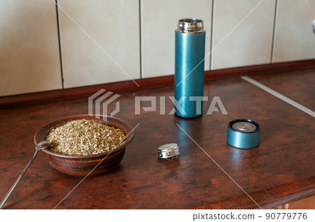 Dried Matricaria chamomilla flowers in a bowl on the kitchen table. An open empty cylindrical thermos with a lid and a teapot next to it. The process of making an herbal tea or infusion of chamomile. Dried Matricaria chamomilla flowers in a bowl on the kitchen table. An open empty cylindrical thermos with a lid and a teapot next to it. The process of making an herbal tea or infusion of chamomile. 90779776