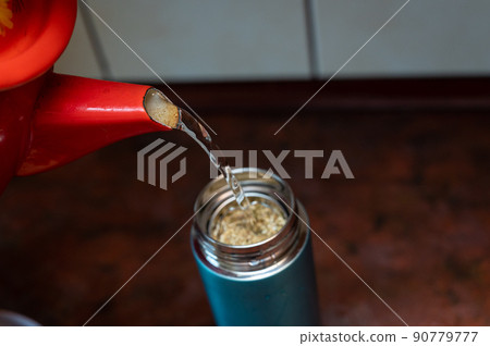 Boiling water from the red chanter is poured into a thermos. The teapot is filled with medicinal herbs. Dried flowers Matricaria chamomilla. Selective focus. Series part. Boiling water from the red chanter is poured into a thermos. The teapot is filled with medicinal herbs. Dried flowers Matricaria chamomilla. Selective focus. Series part. 90779777