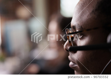 African american operator listening to client on helpline call, using customer service network on headset. Male receptionist working at call center telemarketing assistance. Close up. African american operator listening to client on helpline call, using customer service network on headset. Male receptionist working at call center telemarketing assistance. Close up. 90783332