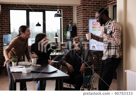 Diverse team of smiling workers and paralyzed man taking break from work in business office. Male employee wheelchair user suffering from physical health condition laughing with colleagues. 90783333