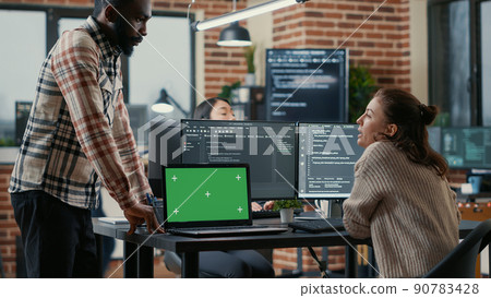 Programer working at desk with multiple computer screens and laptop with green screen chroma key mockup running code. Software developer compiling source algorithm interrupted by colleague. Programer working at desk with multiple computer screens and laptop with green screen chroma key mockup running code. Software developer compiling source algorithm interrupted by colleague. 90783428