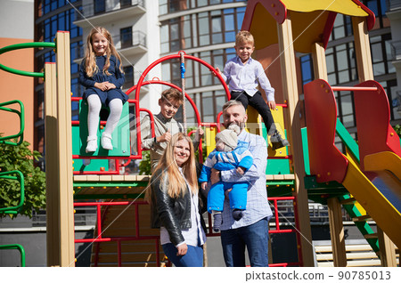 Three cute children hanging out with parents and newborn baby on playground. Front view of happy mum and dad, holding toddler in arms, while other kids smiling at sunny day outdoor. Concept of family. Three cute children hanging out with parents and newborn baby on playground. Front view of happy mum and dad, holding toddler in arms, while other kids smiling at sunny day outdoor. Concept of family. 90785013