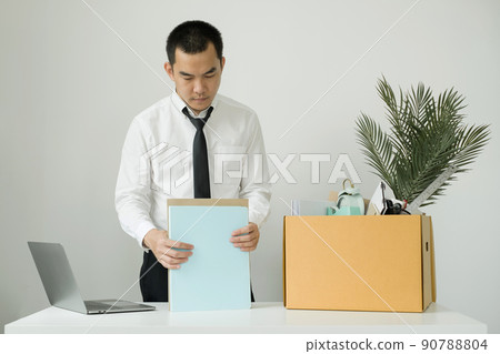 A male office worker is unhappy with being fired from a company packing things into cardboard boxes. The young man was stressed and disappointed by being fired. concept of layoffs and unemployment A male office worker is unhappy with being fired from a company packing things into cardboard boxes. The young man was stressed and disappointed by being fired. concept of layoffs and unemployment 90788804