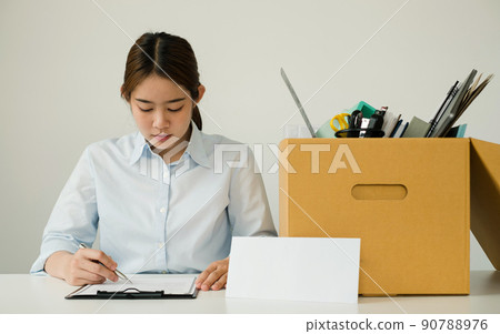A female office worker, dissatisfied with the termination, packs her belongings in a cardboard box and sits down desperately writing her resignation letter. Concept dismissal and unemployment A female office worker, dissatisfied with the termination, packs her belongings in a cardboard box and sits down desperately writing her resignation letter. Concept dismissal and unemployment 90788976