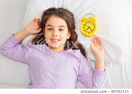 Cute smiling brunette girl lies with a yellow alarm clock on a white pillow Cute smiling brunette girl lies with a yellow alarm clock on a white pillow 90792315