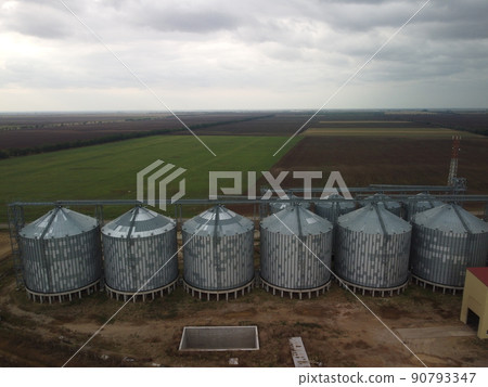 Grain elevator. Metal grain elevator in agricultural zone. Agriculture storage for harvest. Grain elevators on green nature background. Exterior of agricultural factory. Nobody. Grain elevator. Metal grain elevator in agricultural zone. Agriculture storage for harvest. Grain elevators on green nature background. Exterior of agricultural factory. Nobody. 90793347