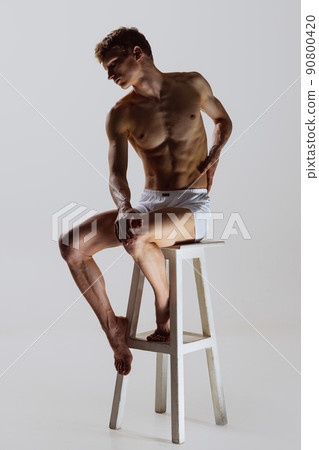 Looks sad, serious. One young shirtless sportive man in white boxer-briefs sitting on high chair isolated on grey background. Art, fashion, diversity, emotions Looks sad, serious. One young shirtless sportive man in white boxer-briefs sitting on high chair isolated on grey background. Art, fashion, diversity, emotions 90800420