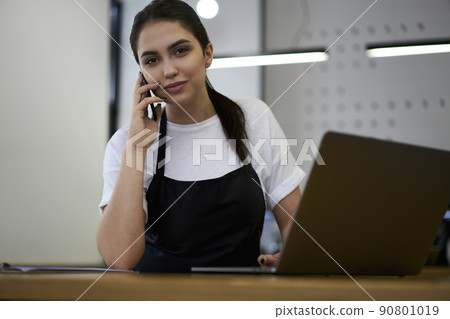 Skilled woman barista making mobile phone talk to service operator checking orders of uniform Skilled woman barista making mobile phone talk to service operator checking orders of uniform 90801019