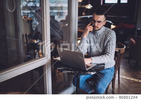 Wistful young handsome man talking on cellphone while doing remote job by using laptop in cafe Wistful young handsome man talking on cellphone while doing remote job by using laptop in cafe 90801204