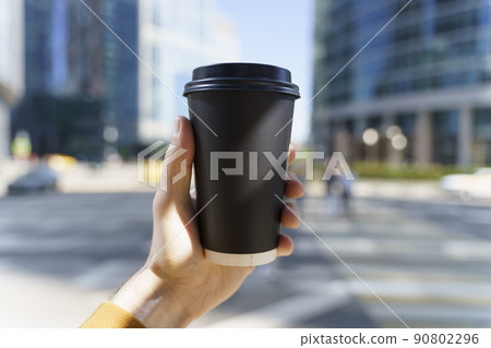Takeaway coffee in a paper cup. A hand with a drink to go on the background of a busy city street. Paper cup mockup 90802296