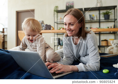 Smiling young mother typing on laptop and doing her online work while her child playing next to her on bed Smiling young mother typing on laptop and doing her online work while her child playing next to her on bed 90805824