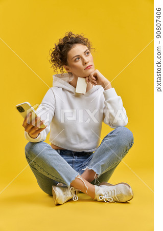 Dreamful caucasian woman in hoodie, with cell phone and bank card sitting on floor. Pensive girl making choice of purchases, before buying, isolated on yellow studio background. Concept of decision. 90807406