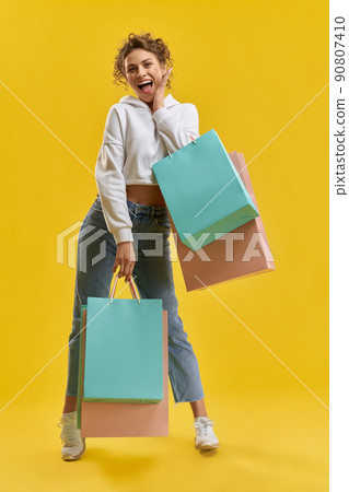 Happy woman in white hoodie holding shopping bags, posing in studio. Front view of pretty model with packages, smiling, shyly holding hand on cheek, isolated on orange background. Concept of shopping. Happy woman in white hoodie holding shopping bags, posing in studio. Front view of pretty model with packages, smiling, shyly holding hand on cheek, isolated on orange background. Concept of shopping. 90807410