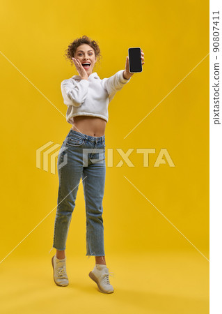 Surprised woman demonstrating blank black screen phone inside. Front view of excited curly girl holding switched off mobile phone, tiptoeing, isolated on orange studio background. Concept of browsing. Surprised woman demonstrating blank black screen phone inside. Front view of excited curly girl holding switched off mobile phone, tiptoeing, isolated on orange studio background. Concept of browsing. 90807411