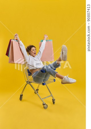 Happy girl holding colorful packages, while riding in shopping cart indoor. Side view of female buyer having fun in shop trolley, while shopping, isolated on orange background. Concept of happiness. Happy girl holding colorful packages, while riding in shopping cart indoor. Side view of female buyer having fun in shop trolley, while shopping, isolated on orange background. Concept of happiness. 90807413