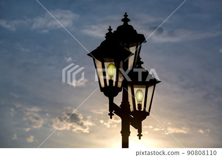 silhouette retro style triple lantern head with transparent glass panels with glow electric light bulbs, iron street lighting in the evening at sunset against a blue sky with sun light. silhouette retro style triple lantern head with transparent glass panels with glow electric light bulbs, iron street lighting in the evening at sunset against a blue sky with sun light. 90808110