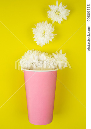 A paper cup for takeaway coffee in pink color lies on a yellow background, white flowers lie out of it. High quality photo 90809518