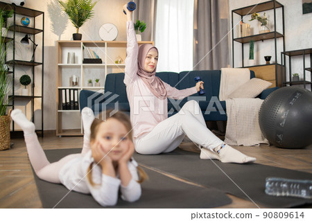 Attractive positive muslim mom doing sports using dumbbells while her cute little girl is resting Attractive positive muslim mom doing sports using dumbbells while her cute little girl is resting 90809614