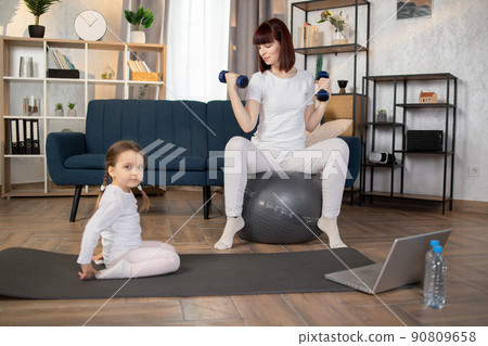 Woman doing sports sitting on fitball with dumbbells in hands while herdaughter sitting on mat 90809658