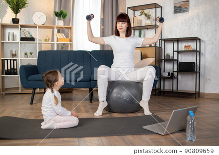 Woman doing sports sitting on fitball with dumbbells in hands while herdaughter sitting on mat 90809659