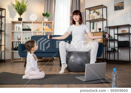 Woman doing sports sitting on fitball with dumbbells in hands while herdaughter sitting on mat Woman doing sports sitting on fitball with dumbbells in hands while herdaughter sitting on mat 90809660