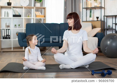 Happy mother with little daughter meditating, sitting in easy seat pose on mat at home Happy mother with little daughter meditating, sitting in easy seat pose on mat at home 90809700