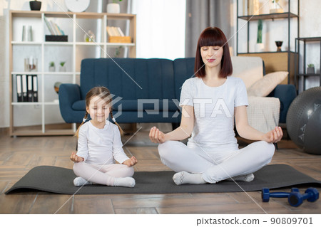 Mother and cute little girl smiling while sitting with closed eyes in lotus pose with on yoga mat Mother and cute little girl smiling while sitting with closed eyes in lotus pose with on yoga mat 90809701