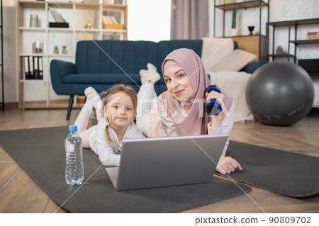 Smiling young muslim mother lying with her cute little daughter on mat on living room floor at home Smiling young muslim mother lying with her cute little daughter on mat on living room floor at home 90809702