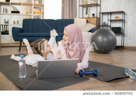 Smiling young muslim mother lying with her cute little daughter on mat on living room floor at home Smiling young muslim mother lying with her cute little daughter on mat on living room floor at home 90809703