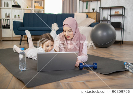 Arabic mum and daughter staring on laptop screen, lying on the mat on floor in living room. 90809706