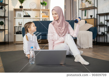 Young muslim mom and daughter doing sports, fitness exercises with laptop on the floor at home. 90809709