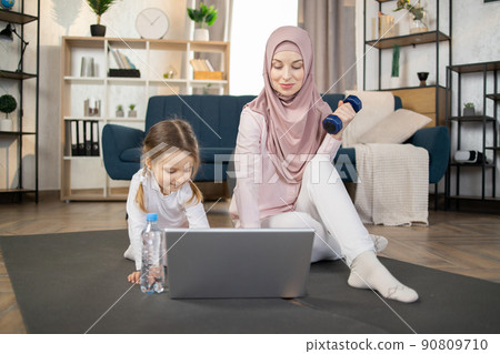 Young muslim woman doing sports gymnastics on the mat at home with her little cute daughter Young muslim woman doing sports gymnastics on the mat at home with her little cute daughter 90809710
