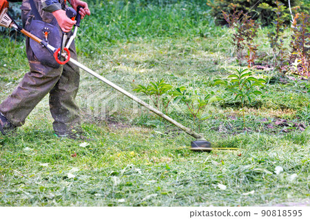 A utility worker mows grass with a petrol trimmer against a green lawn in blur. 90818595