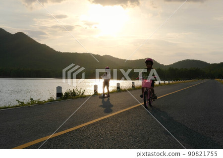 Cute little girl riding a bicycle and her sister riding a scooter on a lake road at sunset. Happy sisters doing outdoor activities together. Healthy Summer Activities for Kids. 90821755